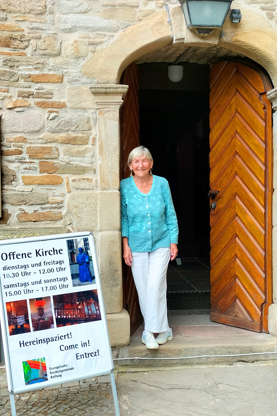 Frau steht vor der geöffneten Kirchentür mit dem Plakat der Öffnungszeiten der offenen Kirche