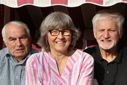 Drei lächelnde ältere Erwachsene posieren zusammen im Freien unter einer gestreiften Markise.