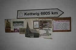 Schild "Kettwig 6805 km" über einer Collage aus Fotos und Papieren.