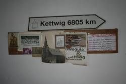 Schild "Kettwig 6805 km" über einer Collage aus Fotos und Papieren.