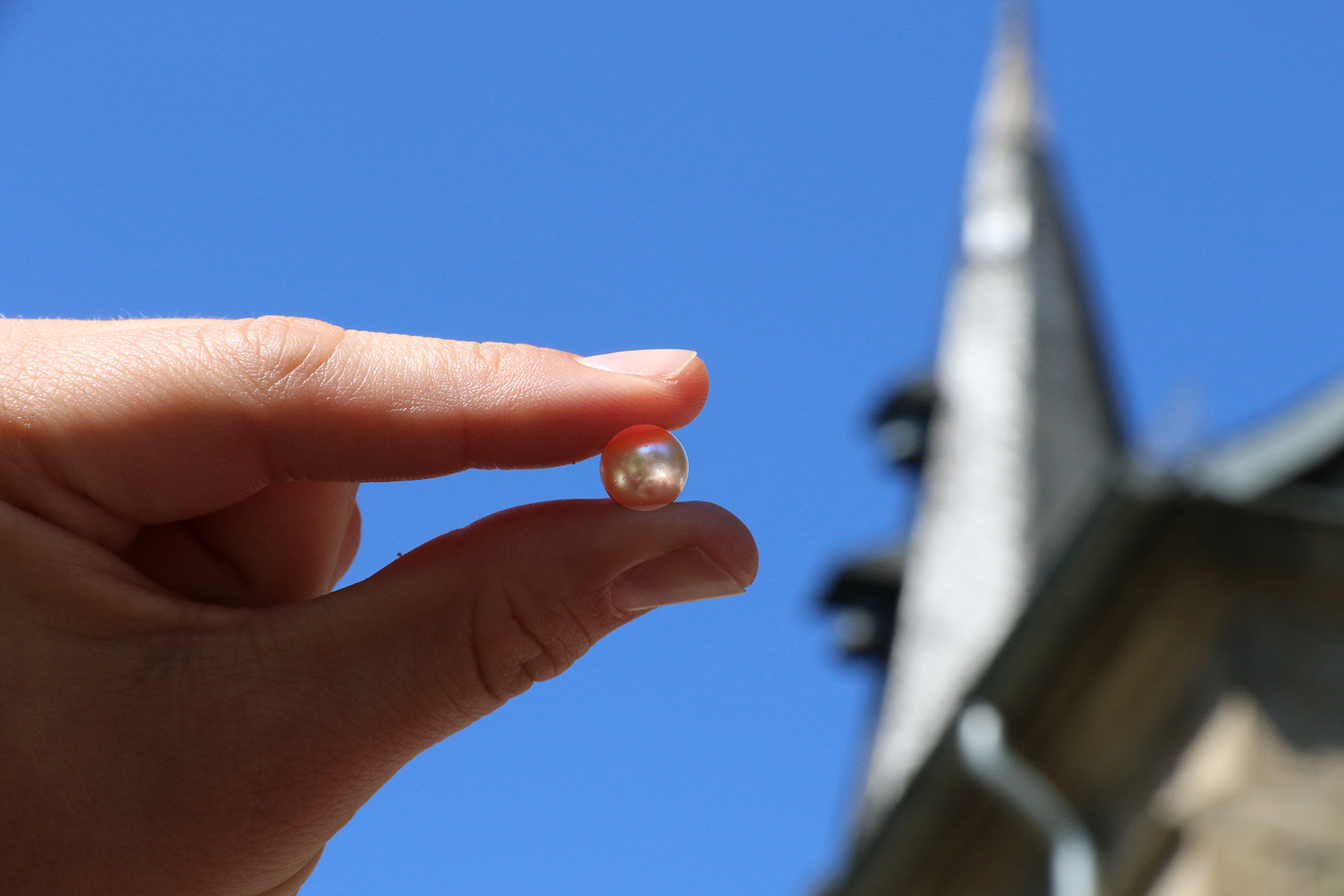 Eine Hand hält eine Perle vor einem Kirchturm.